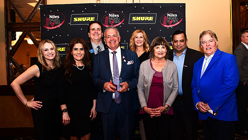 Township officials pose for a picture with honoree, Food Pantry Director Michael Pitzaferro in front of the Niles Chamber of Commerce and Industry&rsquo;s 2026 Night of Roses banner. They are dressed in formal attire and smiling at the camera. Michael is holding the Community Excellence Award.