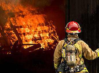 A firefighter wearing a tan turnout coat, yellow reflective stripes, and a red helmet stands with their back to the camera, looking at a large wooden structure engulfed in bright orange flames and smoke.
